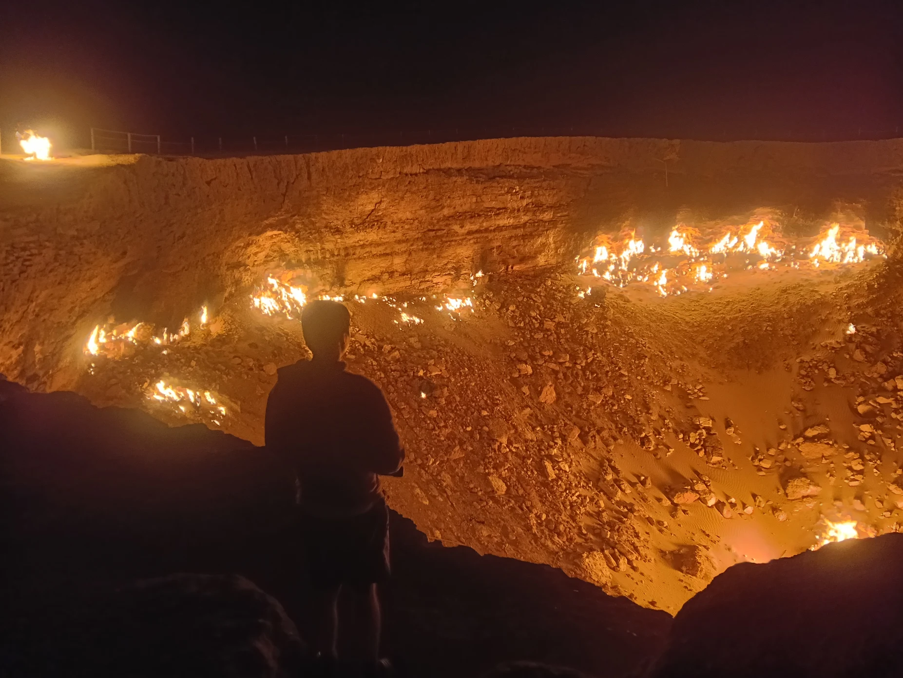 3-Day Tour: Farap ↔ Shavat via Merv, Ashgabat, Darvaza & Kunya-Urgench Traveler standing beside the Darvaza Gas Crater at night as natural gas flames illuminate the desert crater in Turkmenistan