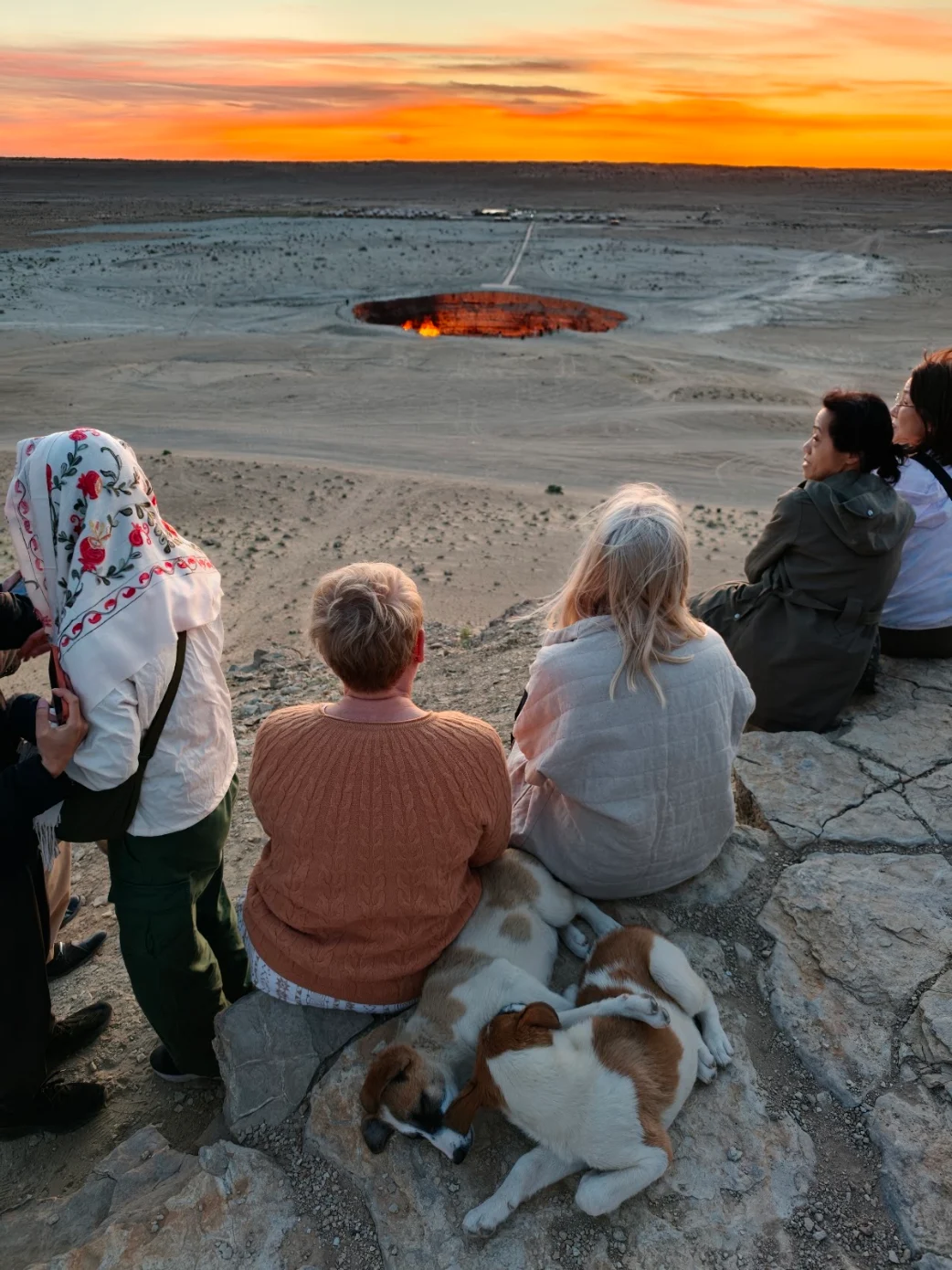 3 Day Turkmenistan Tour from Ashgabat: Darvaza & Night Train to Bukhara International travelers watching sunrise over the Darvaza Gas Crater in the Karakum Desert, with two dogs resting beside them in Turkmenistan