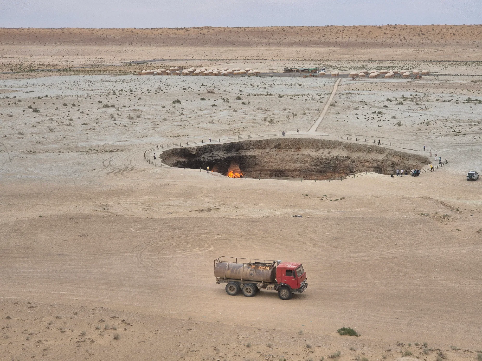 Daytime panoramic view of Darvaza Gas Crater with visitors, a truck crossing the desert and the yurt camp visible in the distance