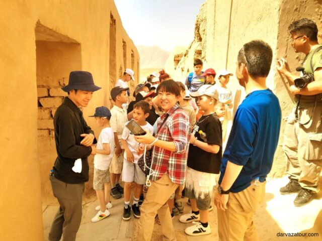 A tourist group with children who came to travel to Nissa explores ancient ruins in Turkmenistan. Visitors wearing protective headgear examine the historical site under warm sunlight.