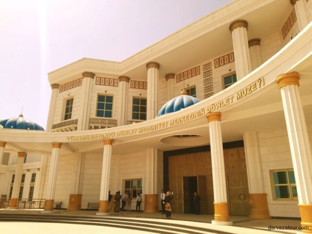 Exterior of the National Museum of Turkmenistan featuring beige architecture with blue domes, prominent columns at the entrance, and decorative inscriptions above the main doorway. The grand building displays traditional Turkmen architectural elements with visitors visible at the entrance.