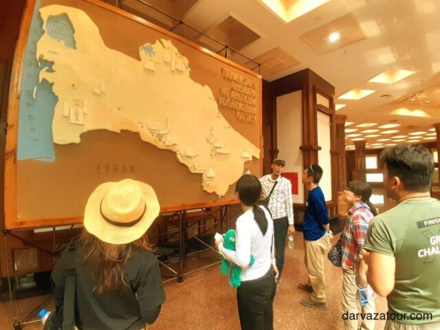 Tour group examining a large wall map of Turkmenistan in a museum. Visitors stand in front of a beige relief map of the country with the blue Caspian Sea on the left. The guide explains Turkmenistan's geography while tourists, including a person in a straw hat, listen attentively.