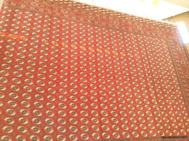 The largest carpet in the world in Turkmenistan featuring a rich red background with ornate gold geometric patterns and traditional motifs, displayed in a museum setting