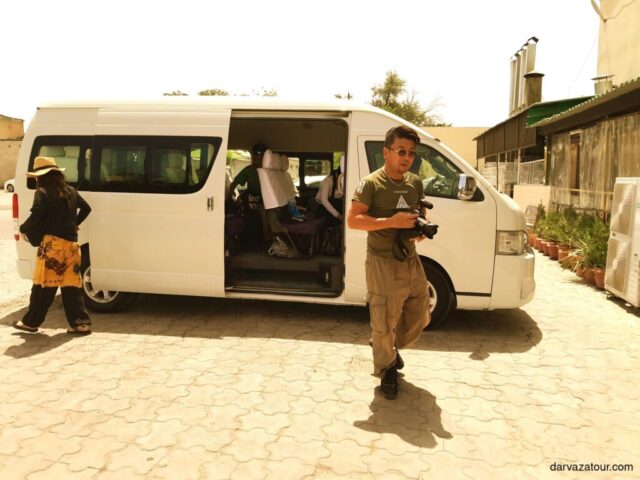 White Toyota Hiace tour van with open sliding door parked on a paved street in Turkmenistan. A tour guide with a camera stands in front of the vehicle while a person in traditional dress approaches the van.