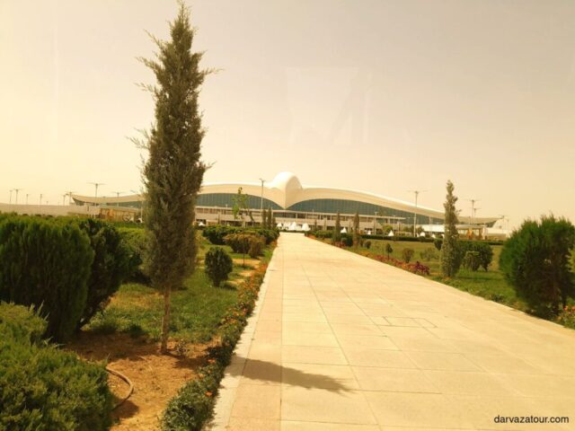 Access road to Ashgabat International Airport with picturesque greenery on both sides. The modern airport building with its distinctive architecture is visible in the background in the rays of the setting sun.