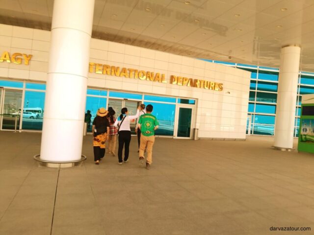 Tourists heading to the international departures area of Ashgabat Airport in Turkmenistan. A golden sign "INTERNATIONAL DEPARTURES" can be seen on the wall.