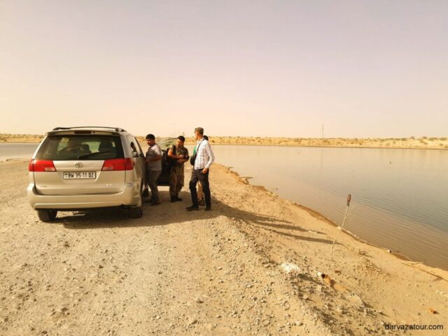 Tourists stopped at a lake on the way to the Darvaza crater. A silver Toyota minivan is parked on the sandy shore, and a group of travelers has gathered next to it to enjoy the view of the water body in the desert landscape of Turkmenistan.