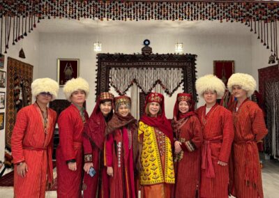 Group of Singaporean tourists wearing traditional Turkmen national costumes inside the Carpet Museum in Ashgabat, smiling and posing together in front of a decorative Turkmen carpet background.