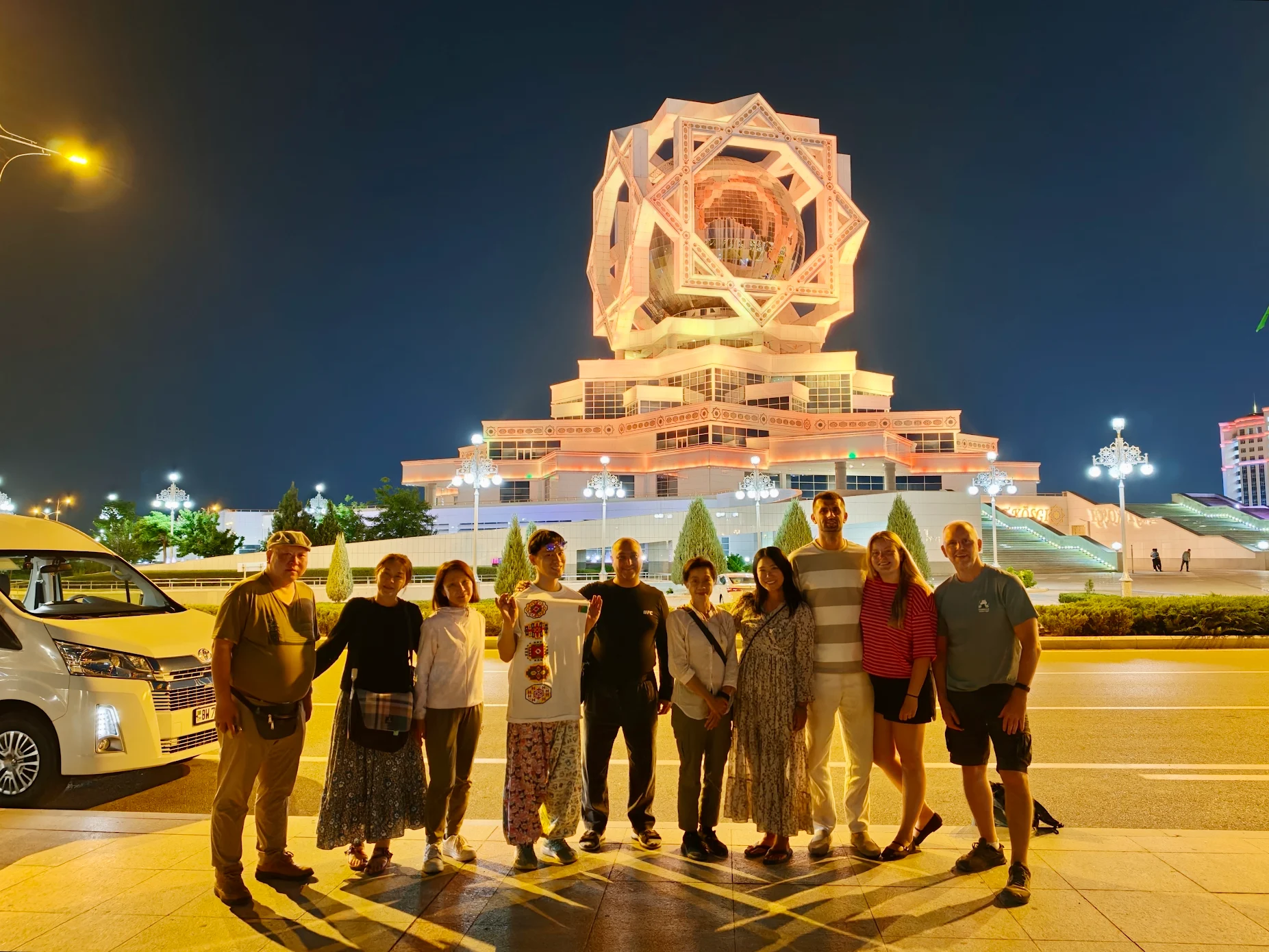 Tour Group at Alem Center in Ashgabat International tour group with local guide Yusup posing beside a Toyota Hiace in front of the illuminated Alem Cultural and Entertainment Center in Ashgabat, Turkmenistan