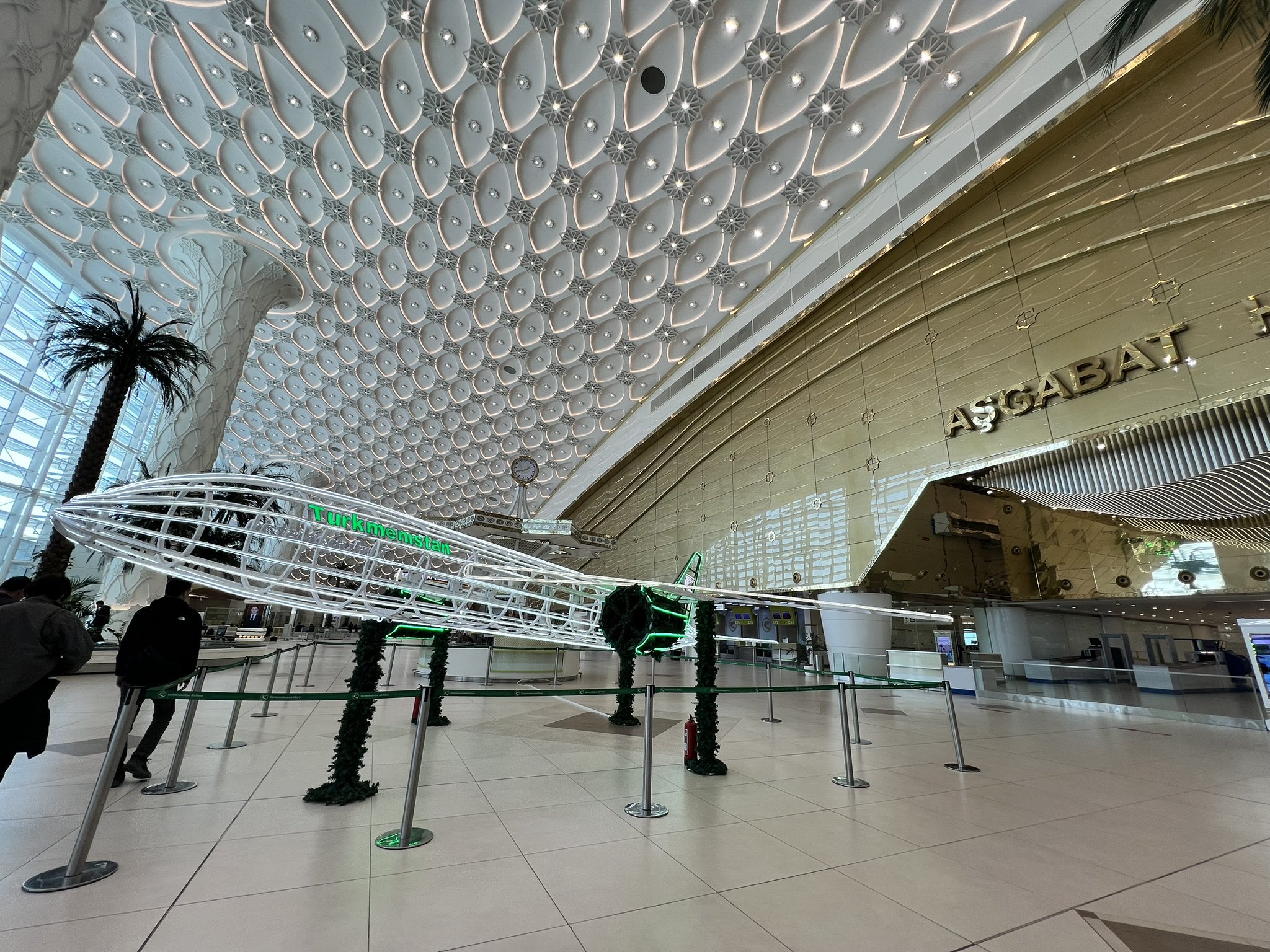 Interior of Ashgabat International Airport with golden walls, modern ceiling design, and a Turkmenistan Airlines airplane model.