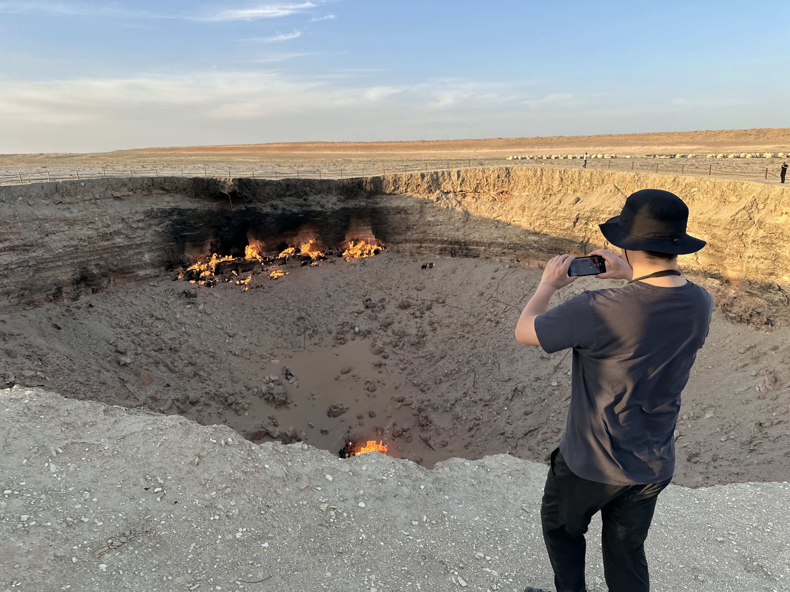 Japanese Traveler Photographing Darvaza Gas Crater Japanese traveler wearing a sun hat and taking a photo of Darvaza Gas Crater with the desert yurt camp in the background