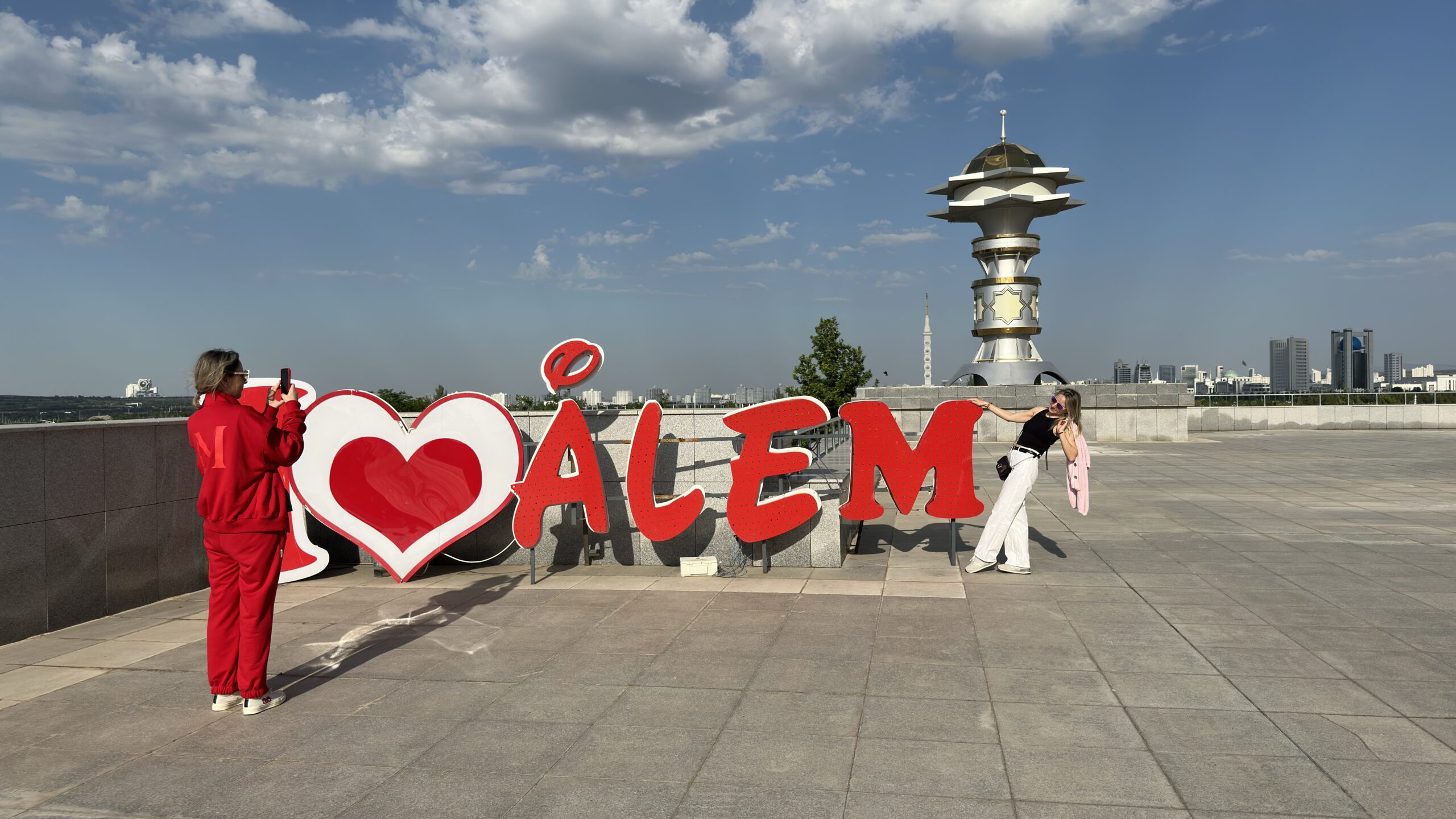 Two Russian travelers taking photos beside the I Love Alem sign at a panoramic viewpoint overlooking Ashgabat, Turkmenistan