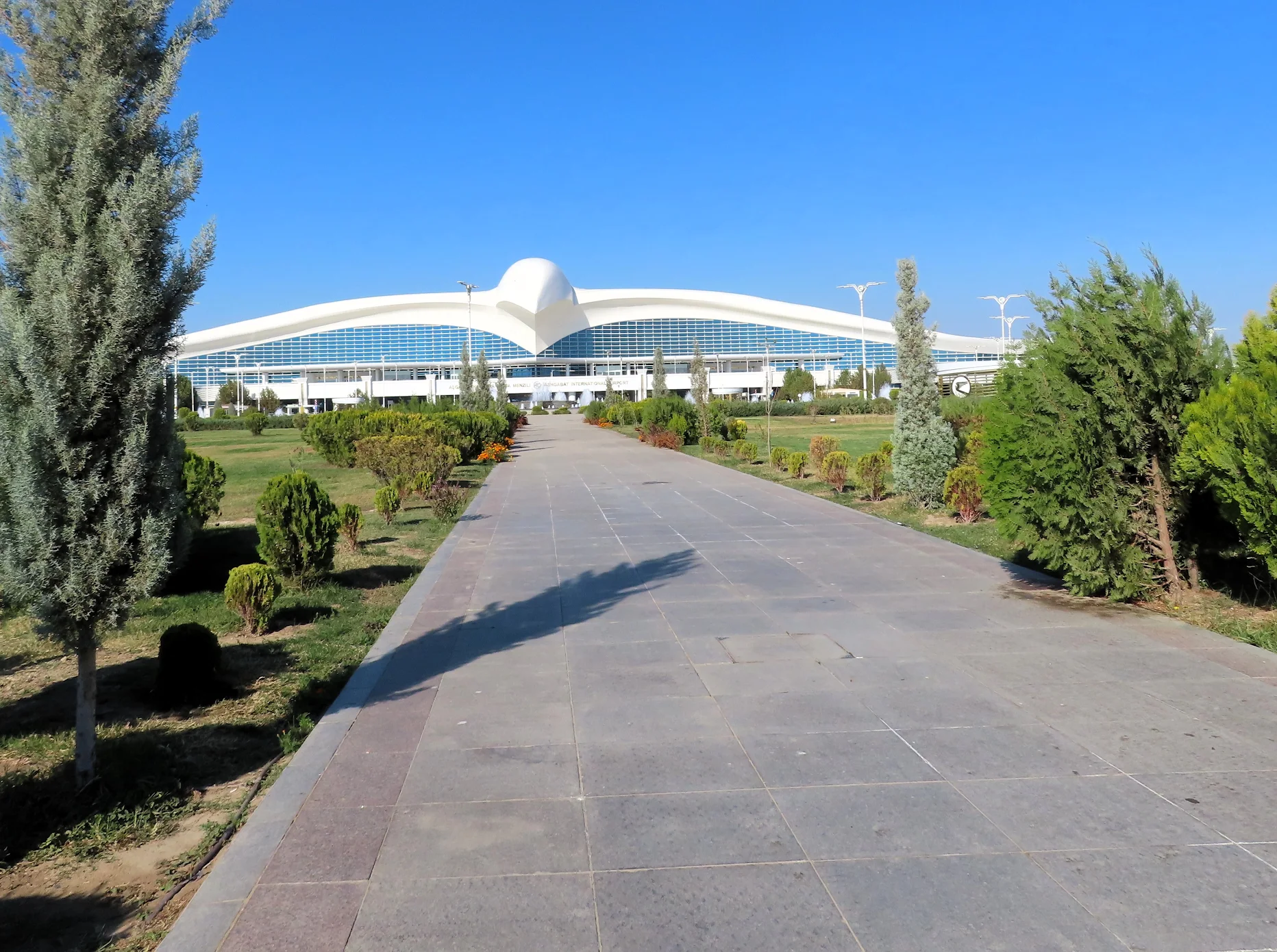 Ashgabat International Airport with its distinctive white falcon-shaped terminal surrounded by landscaped gardens under a clear blue sky.