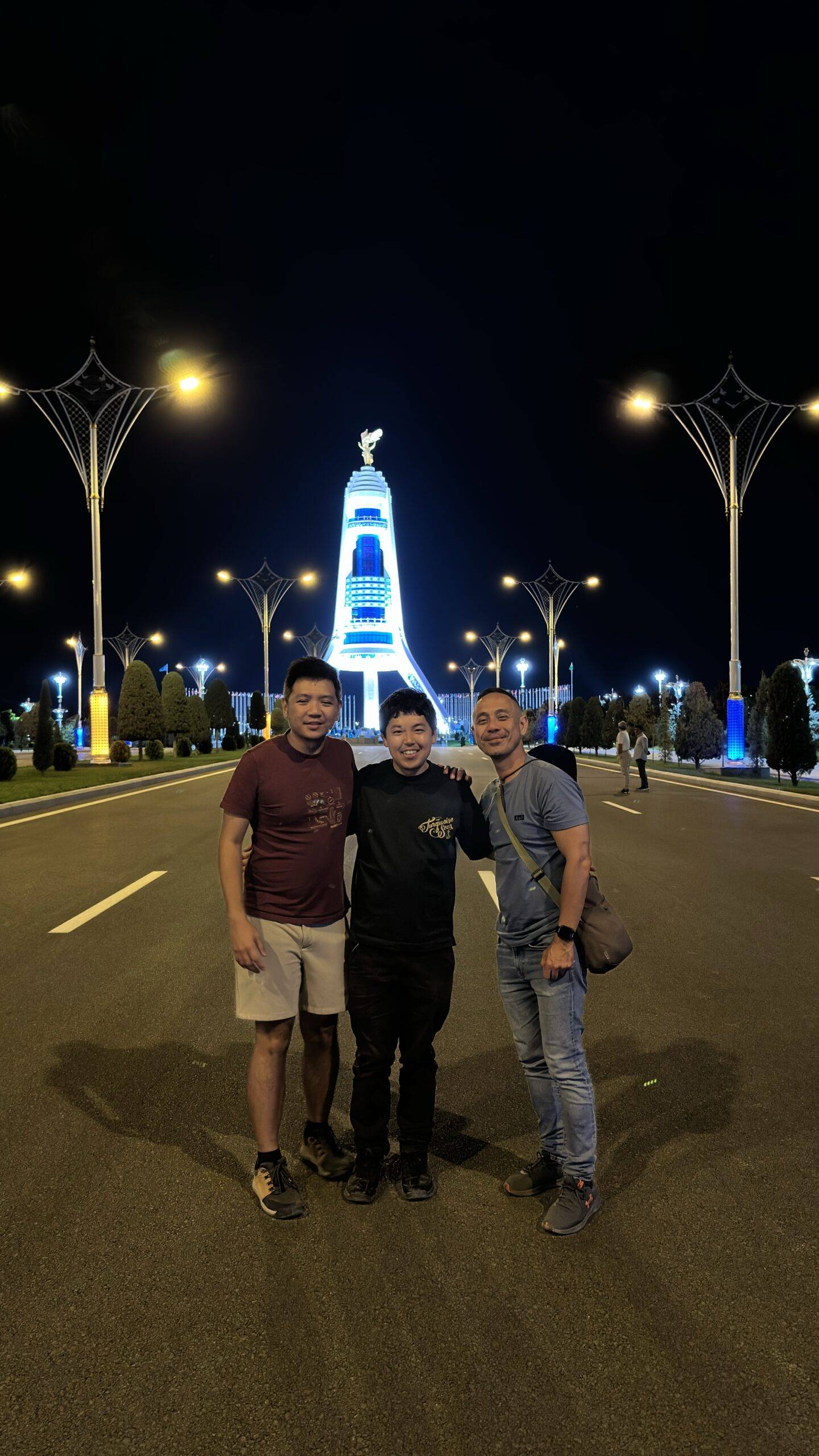 Three Malaysian travelers posing in front of the illuminated Monument of Neutrality at night in Ashgabat, Turkmenistan