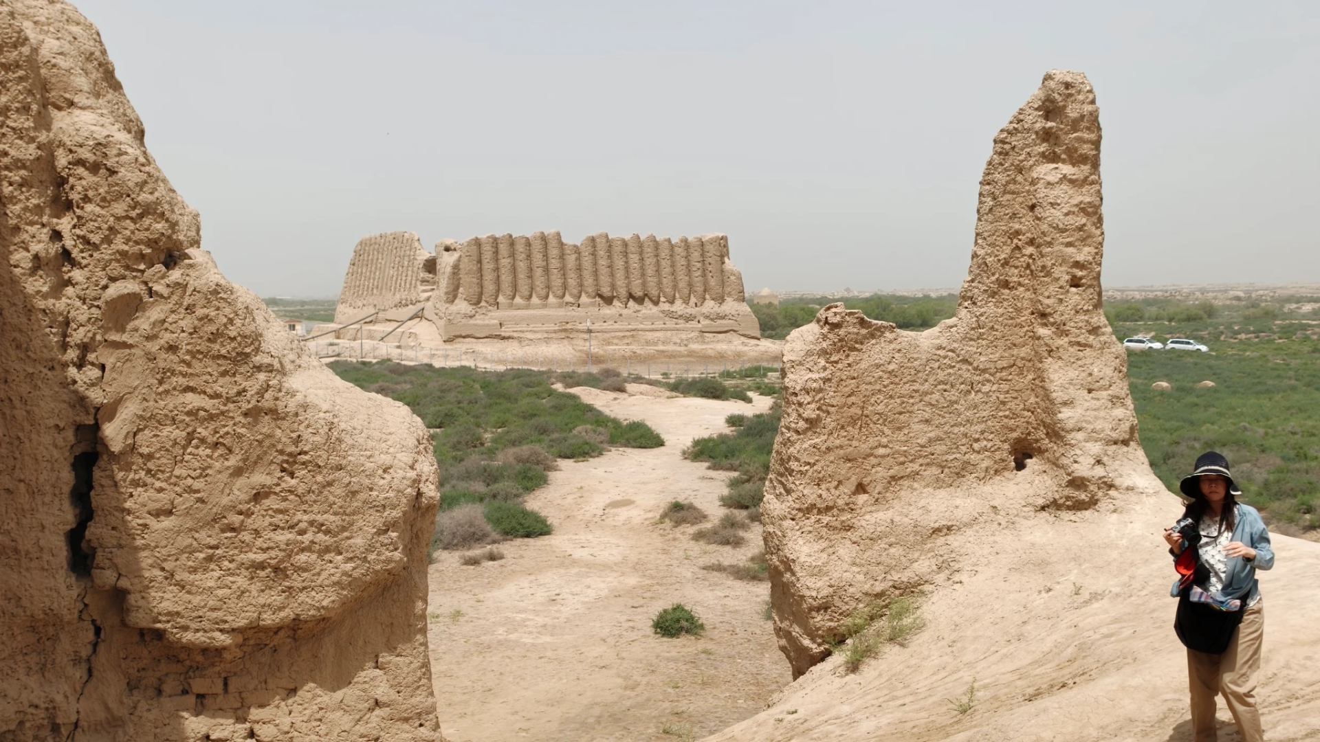 Traveler exploring ancient mud-brick ruins with the distinctive corrugated walls of Great Kyz Kala visible in the distance at Ancient Merv.