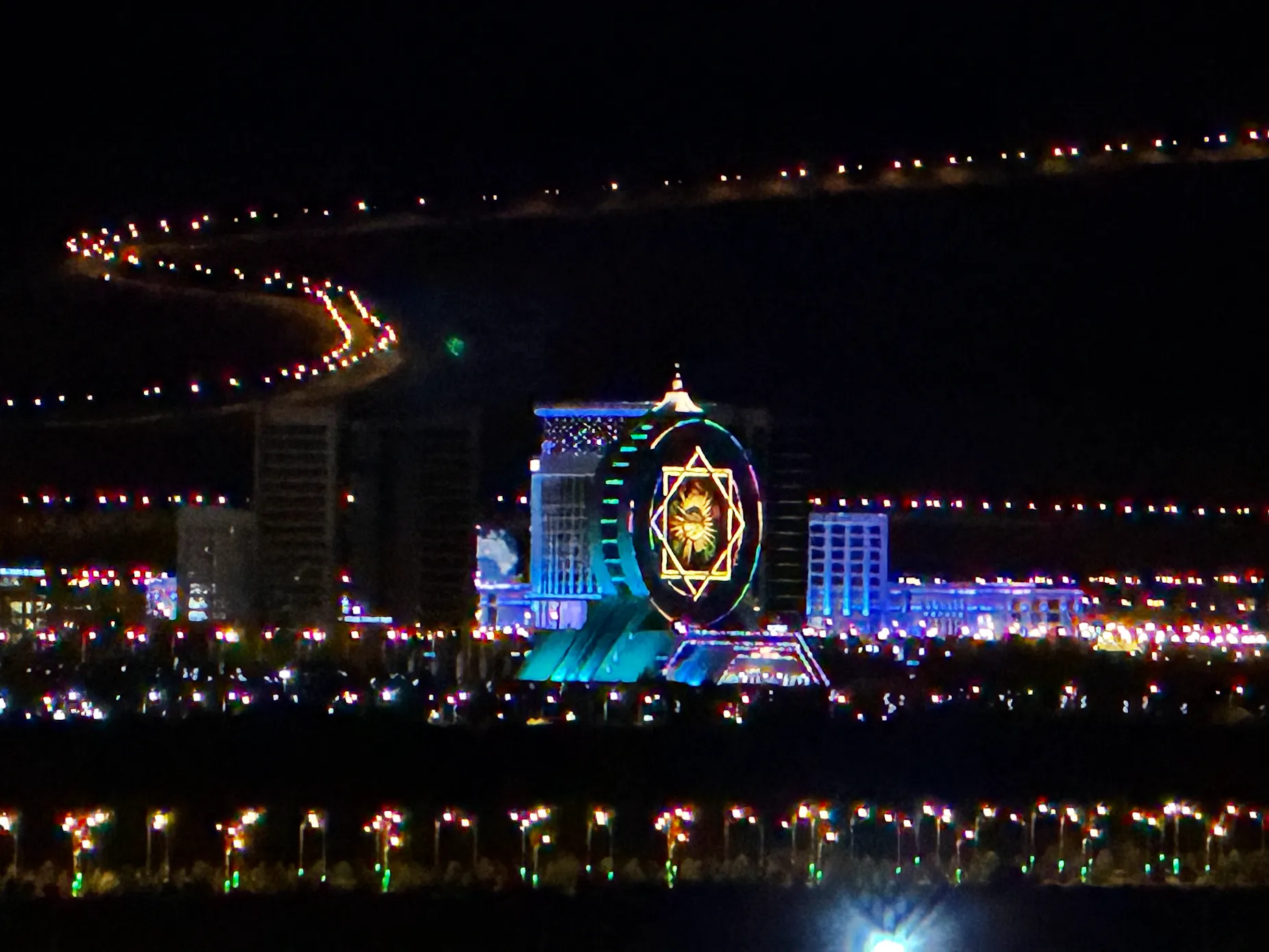 Colorfully illuminated modern architecture and city lights of Ashgabat glowing at night against the dark foothills of the Kopet Dag Mountains.